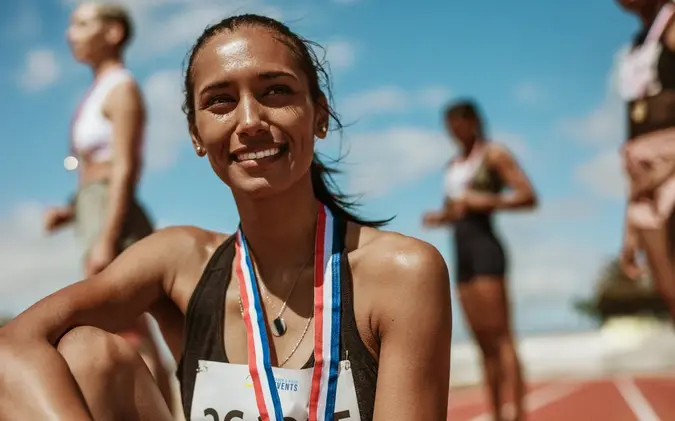 Female athlete with medal sitting on racetrack with team standing in background.